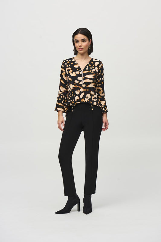 A woman with shoulder-length dark hair is standing against a plain backdrop. She is wearing the Silky Knit Animal Print Tunic by Joseph Ribkoff, paired with black high-waisted trousers that have gold buttons on the front and black heeled boots.