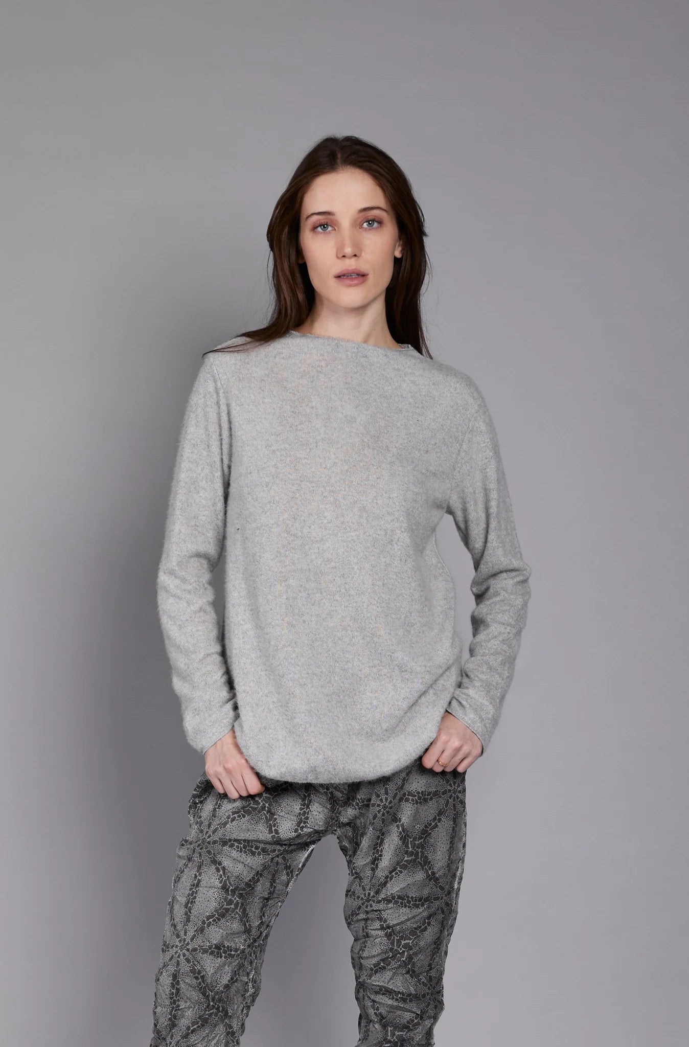 A person with long hair is standing against a plain gray background. They are wearing the JY4138 TOP by ELISSIA, made of soft breathable fabric, and patterned gray pants, with their left hand resting in their pants pocket and their right arm hanging by their side.