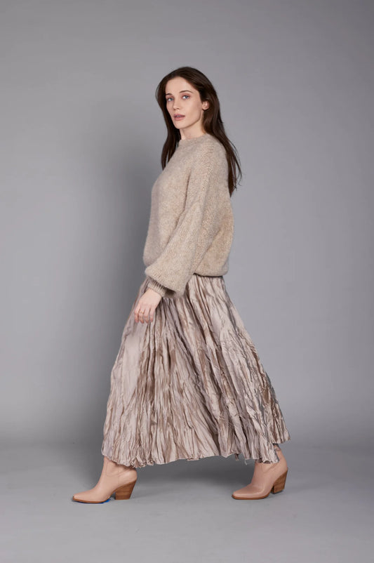 A person with long brown hair wearing an ELISSIA chic beige, oversized sweater (EL3565 TOP) and a gray, pleated, ankle-length skirt is walking sideways. They are also wearing brown ankle boots with a low heel. The background is a plain gray studio setting.