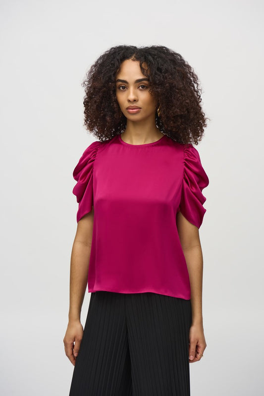 A woman with curly hair, wearing the Joseph Ribkoff Satin Straight Top With Shirred Sleeves (style number 244023) and black pleated pants, stands against a plain white background. She gazes directly at the camera with a neutral expression.