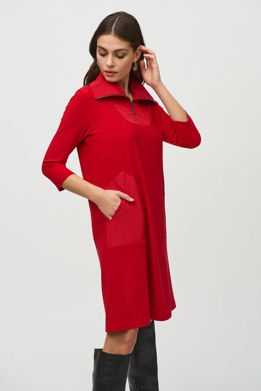 A woman with long dark hair wears the Silky Knit And Memory Straight Dress 244045 by Joseph Ribkoff, a knee-length dress with an A-line silhouette in red bouclé mesh fabric. The dress features a zipped neckline and quarter sleeves. She stands against a plain background, one hand raised to her head as she looks down to the left. Black knee-high boots complete her look.