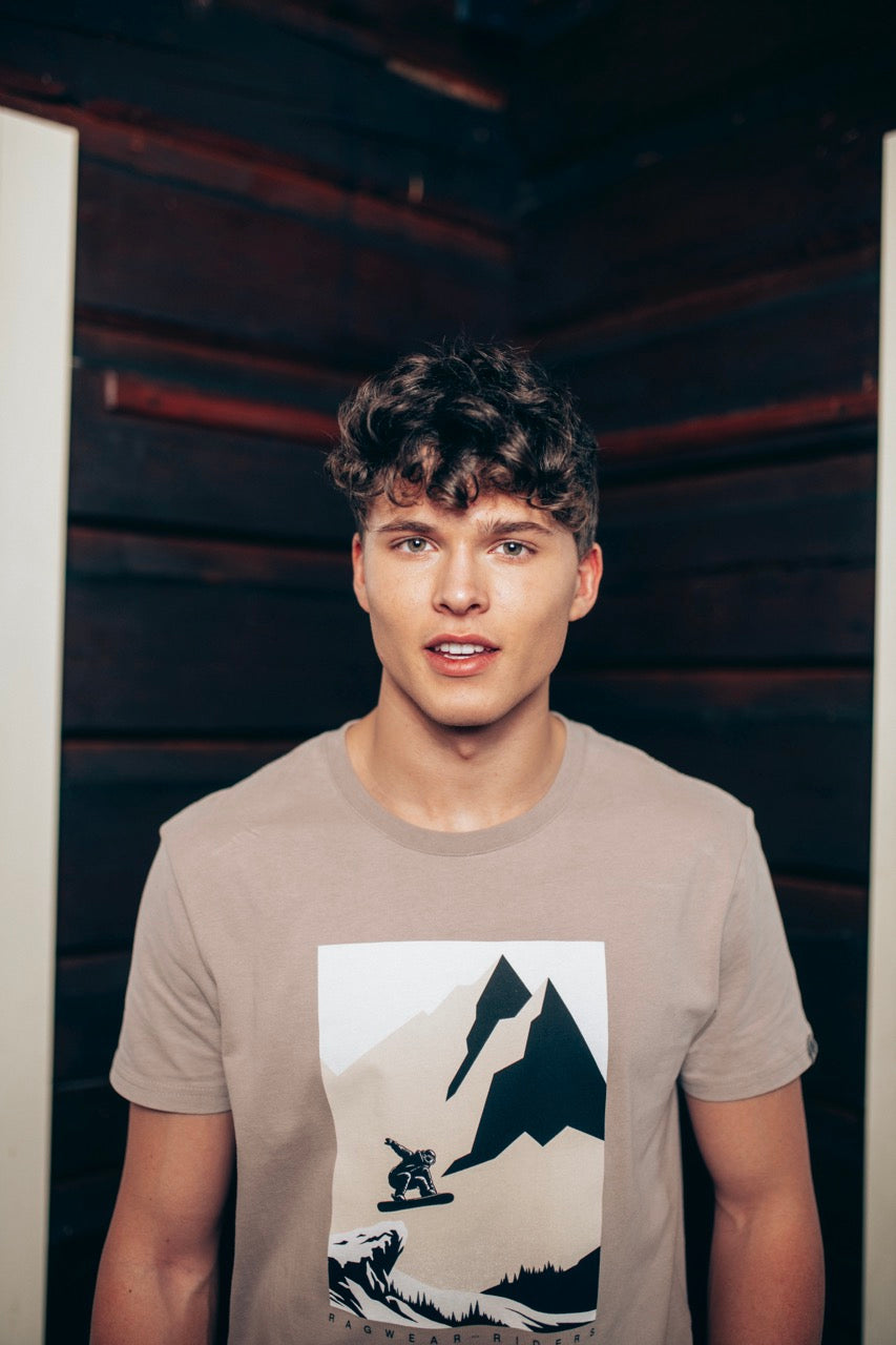 A man with short hair and stubble is wearing the Roggero T-Shirt 2422-15024-7001 from RAGWEAR, a beige, eco-friendly shirt featuring a mountain and bear graphic on the front. He pairs it with blue jeans and stands against a plain white background, looking directly at the camera with a neutral expression.