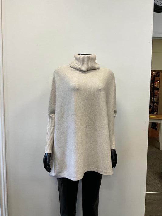 A mannequin dressed in an ETERNELLE brand "Tunic with Turtle Neck Beige" sweater, made in Italy from premium fabrics, and black pants is displayed against a white wall in a retail store. The light gray long-sleeve turtleneck tunic outfit appears casual and cozy.