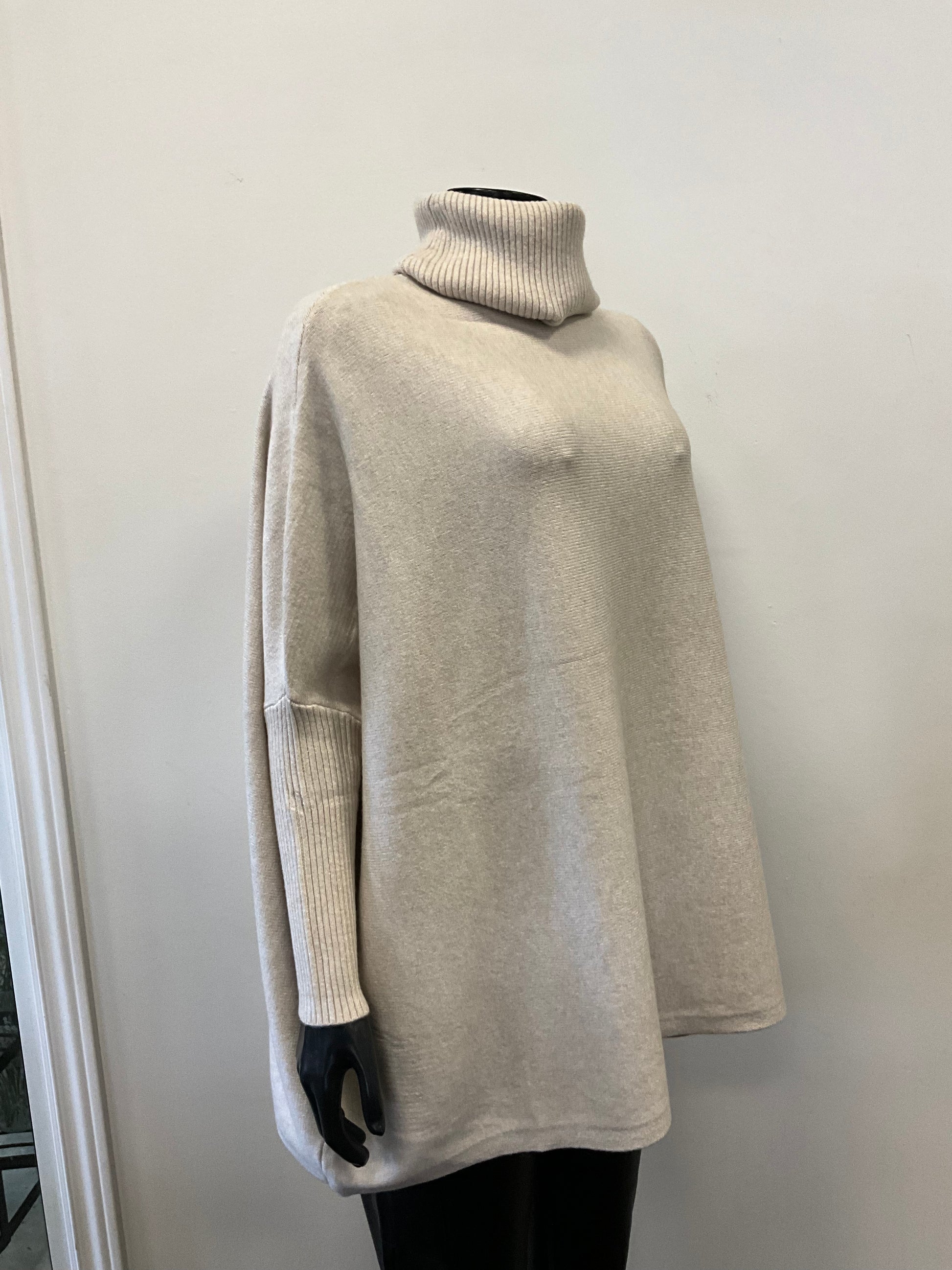 A mannequin dressed in an ETERNELLE brand "Tunic with Turtle Neck Beige" sweater, made in Italy from premium fabrics, and black pants is displayed against a white wall in a retail store. The light gray long-sleeve turtleneck tunic outfit appears casual and cozy.