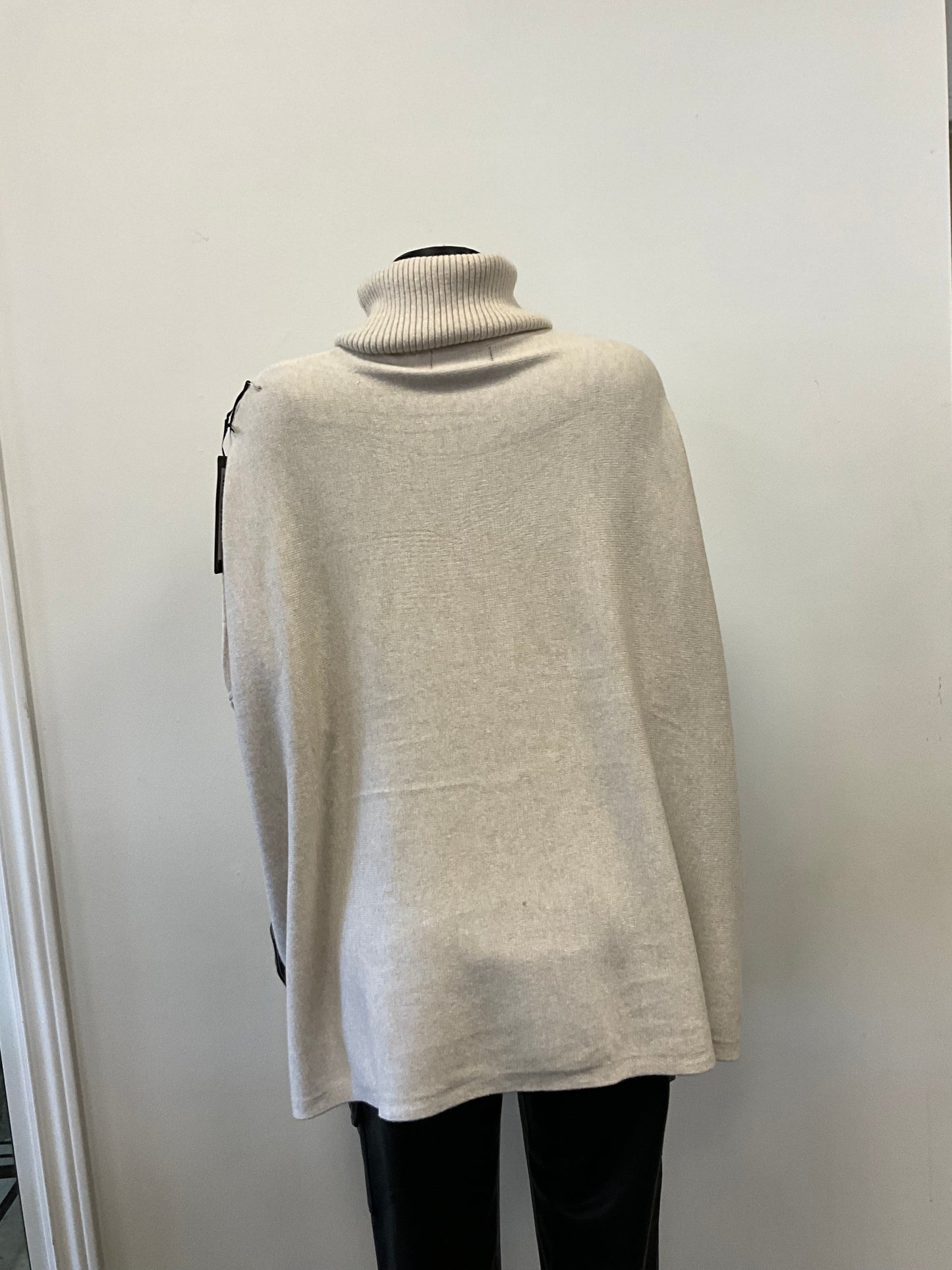 A mannequin dressed in an ETERNELLE brand "Tunic with Turtle Neck Beige" sweater, made in Italy from premium fabrics, and black pants is displayed against a white wall in a retail store. The light gray long-sleeve turtleneck tunic outfit appears casual and cozy.