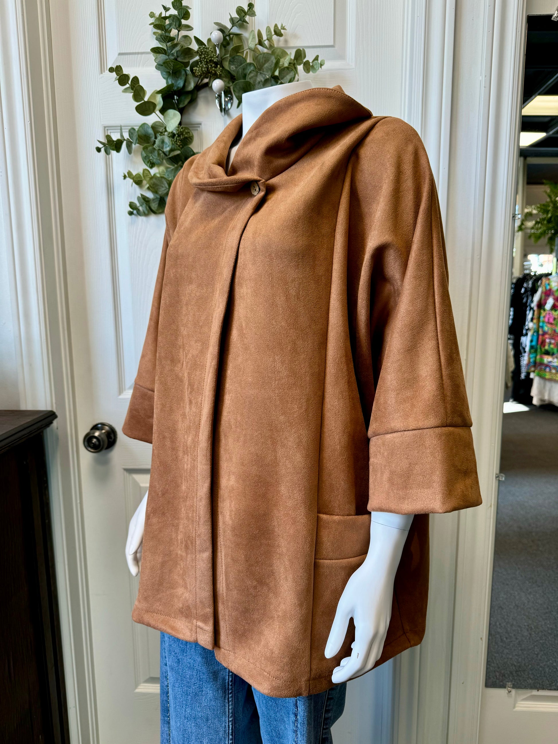 A mannequin showcases the ELISSIA BC12762 Shawl 12762 - Elissia, a camel-colored, suede-like jacket featuring wide sleeves and a distinctive buttoned collar. This luxurious jacket, complete with two front pockets, is elegantly paired with blue jeans. In the background, an assortment of clothing items and some greenery are hanging on a door.
