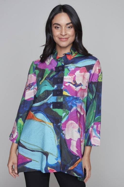 A woman with long black hair wears a colorful, patterned Clare Desjardins Woven Top Tunic 91711 featuring vibrant shades of blue, pink, purple, and green. The tunic has a button-down front and three-quarter length sleeves. She stands against a grey background, smiling with her hands relaxed at her sides.