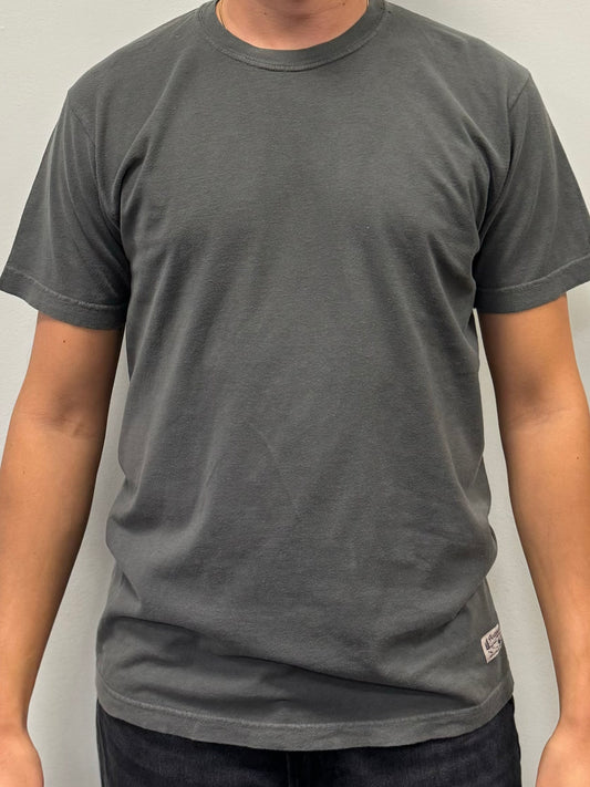 A person wearing a plain, dark grey Northbound Supply Co T-shirt and dark pants stands against a light grey background. The 100% cotton T-shirt from Northbound Supply Co features short sleeves and a small tag at the bottom on the left side. The person's hands are by their sides, and their head is not visible in the image.