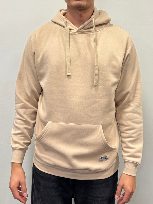 A person is wearing the Hoodie from Northbound Supply Co in beige, featuring a hood with drawstrings and a front kangaroo pocket. The sweatshirt includes a small rectangular tag at the bottom. The individual also has on black pants. The off-white background adds to the casual Canadian vibe.