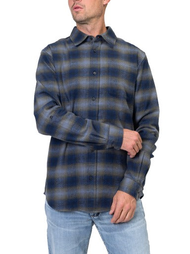 A person is wearing a timeless piece: a Men’s Navy Plaid Flannel Shirt from Silver Jeans Co. paired with light blue jeans. Their right arm is slightly bent, and their left hand is adjusting their right sleeve against the plain white background.