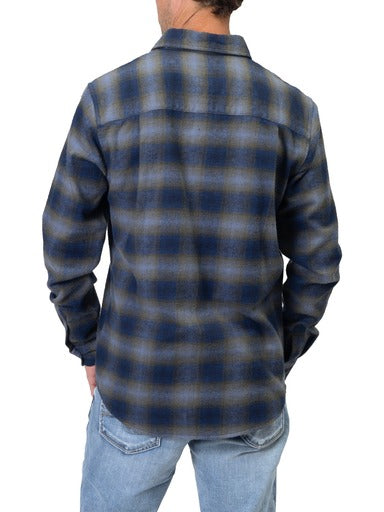 A person is wearing a timeless piece: a Men’s Navy Plaid Flannel Shirt from Silver Jeans Co. paired with light blue jeans. Their right arm is slightly bent, and their left hand is adjusting their right sleeve against the plain white background.