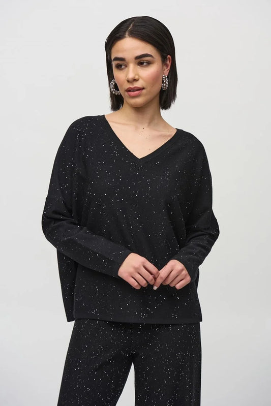 A person with shoulder-length dark hair is wearing the Joseph Ribkoff Sequined Sweater Knit Boxy Top-244921, paired with matching black pants featuring a white speckled pattern. Their hands are clasped together, and they are looking slightly to the side while standing against a plain, light-colored background.