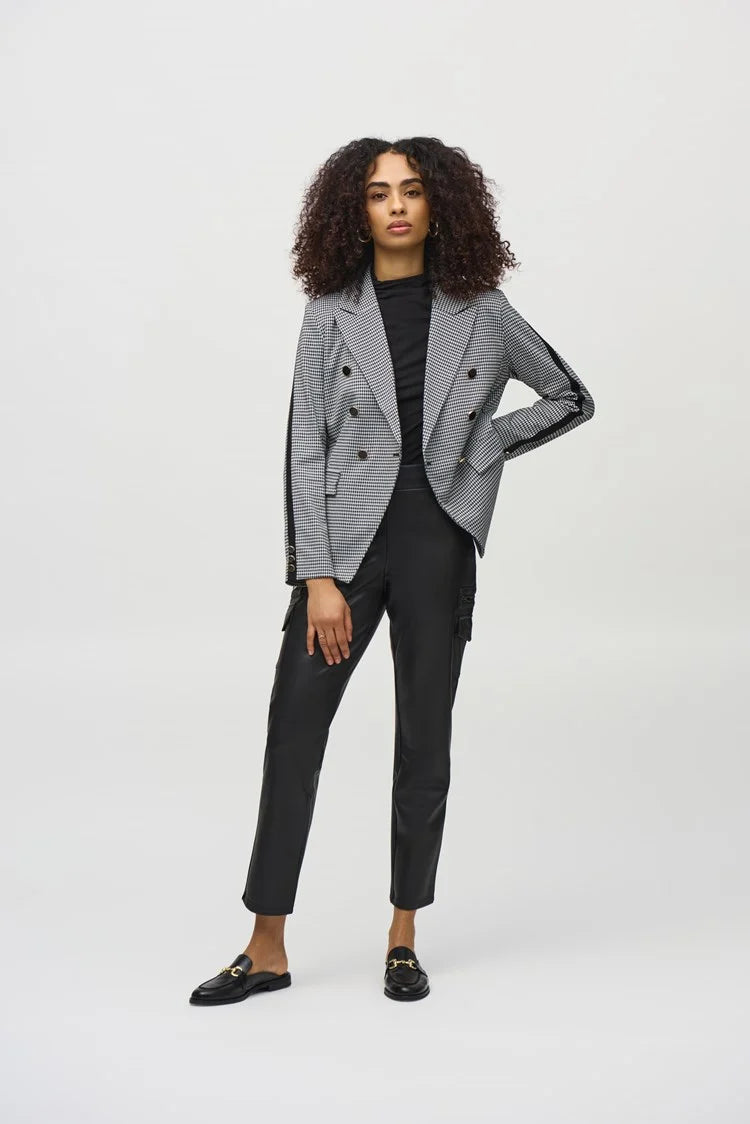 A woman with blonde curly hair is leaning against a wood-paneled wall and a black marble fireplace. She is wearing a houndstooth blazer over a black top and the Joseph Ribkoff Leatherette Slim Fit Pull-On Pants 244939, paired with black loafers. Her expression is contemplative as she poses with one hand on her chin.