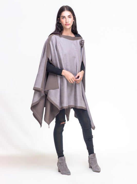 A woman with long dark hair, wearing a light grey Lino Poncho by CHAKEN with an asymmetrical hem and subtle floral accents, stands against a plain white background. She is dressed in black skinny jeans, a black top, and grey ankle boots. Her hands are lightly clasped in front of her.