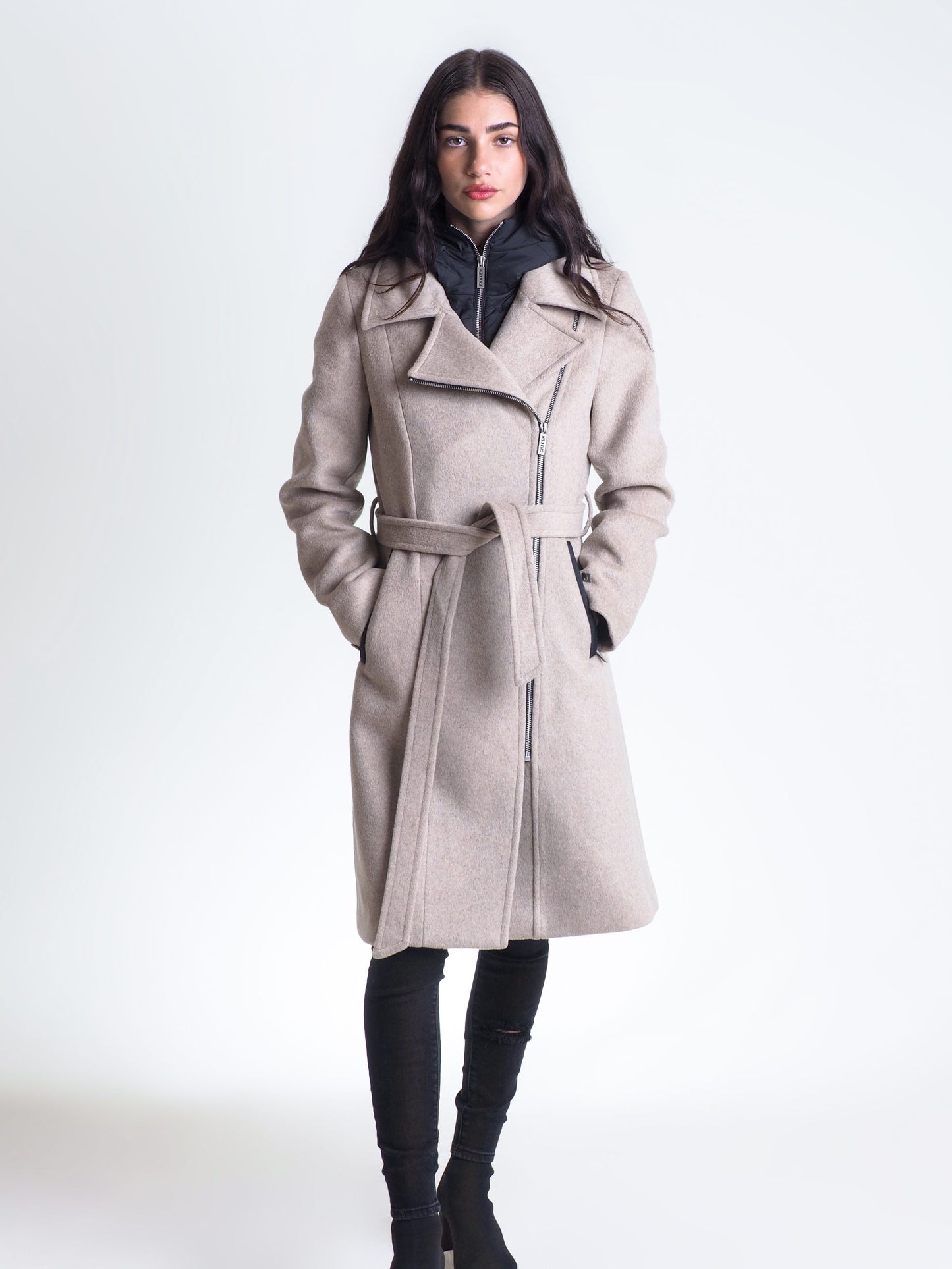 A woman with long dark hair stands against a plain background wearing a beige Luna Coat - CHAKEN with a black inner lining, leggings, and black boots. With her hands in the coat pockets, she looks directly at the camera showcasing CHAKEN's elegance.