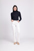 A person with long dark hair stands against a plain light background, wearing a FOIL Fine Merino Polo Neck Top-Navy paired with high-waisted white pants. Their hands are in their pockets, and they are wearing beige shoes. They are smiling gently while facing the camera.