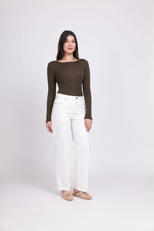 A woman with long dark hair stands against a plain white background. She is wearing the FOIL Fine Merino Crew Neck Top in khaki marl green, paired with high-waisted white jeans and beige flat shoes. She is smiling and looking slightly to the side.