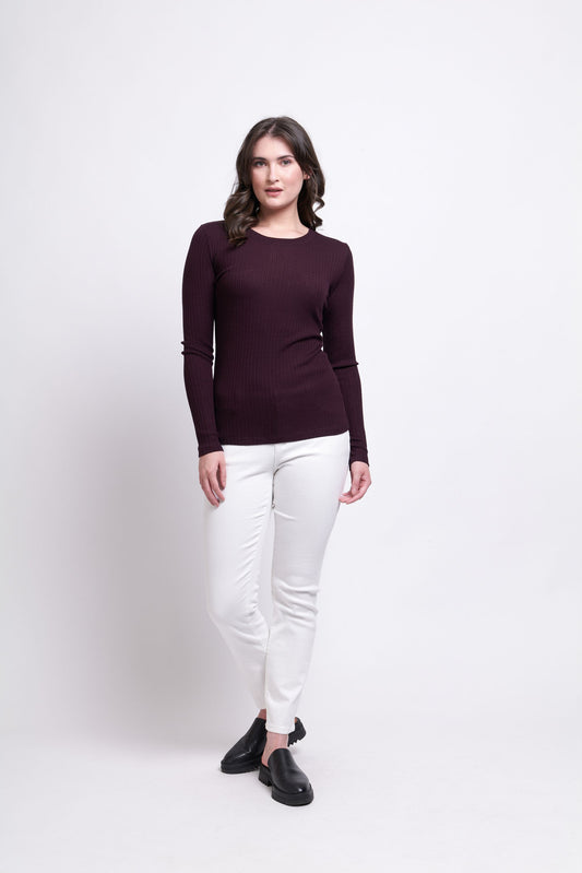 A woman with shoulder-length hair stands against a plain white background, wearing the Fine Merino Crew Neck Top-Plum by FOIL, along with white pants and black shoes. She has a relaxed posture, with one foot slightly in front of the other and her hands by her sides.