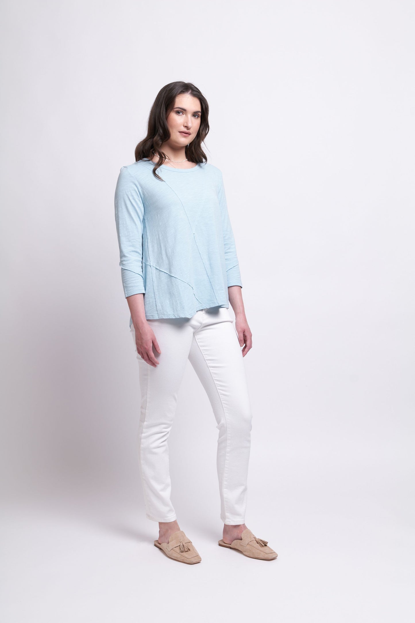 A woman stands against a plain white background, donning the "Seam It All Top" in Fog Blue by FOIL. Pairing this chic, light blue long-sleeved top featuring a subtle triangle pattern with white slim-fit pants and light beige slip-on shoes, she looks straight ahead with her hands relaxed at her sides, showcasing her sophisticated wardrobe choice.