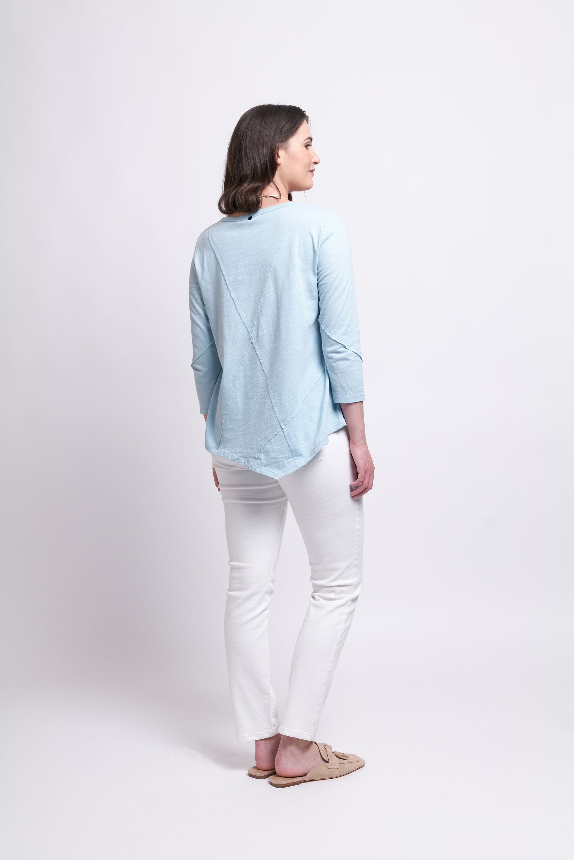 A woman stands against a plain white background, donning the "Seam It All Top" in Fog Blue by FOIL. Pairing this chic, light blue long-sleeved top featuring a subtle triangle pattern with white slim-fit pants and light beige slip-on shoes, she looks straight ahead with her hands relaxed at her sides, showcasing her sophisticated wardrobe choice.