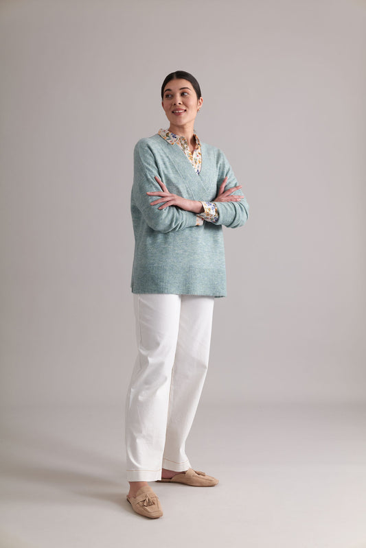 A person with long hair tied back is standing with arms crossed, looking away. They are wearing the light blue, stylish "Come Together Sweater" by FOIL over a floral shirt, white pants, and brown loafers. The background is plain and light-colored.