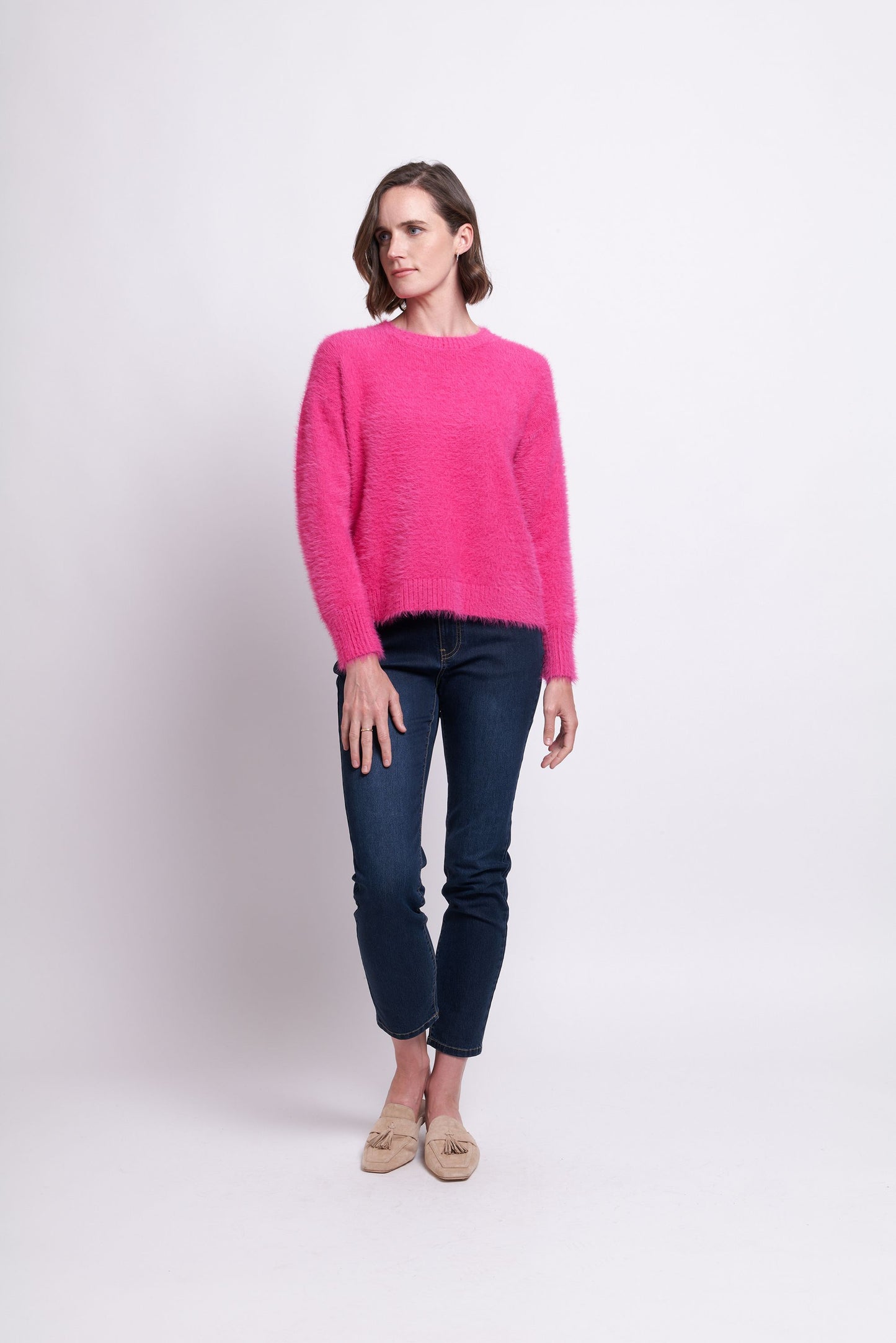 A person with short hair is standing against a plain background, wearing a bright pink, fuzzy "Fluff Love Sweater - FOIL" by FOIL, dark blue jeans, and beige slip-on shoes. The person has one arm relaxed at their side and the other slightly bent with their hand near their hip.