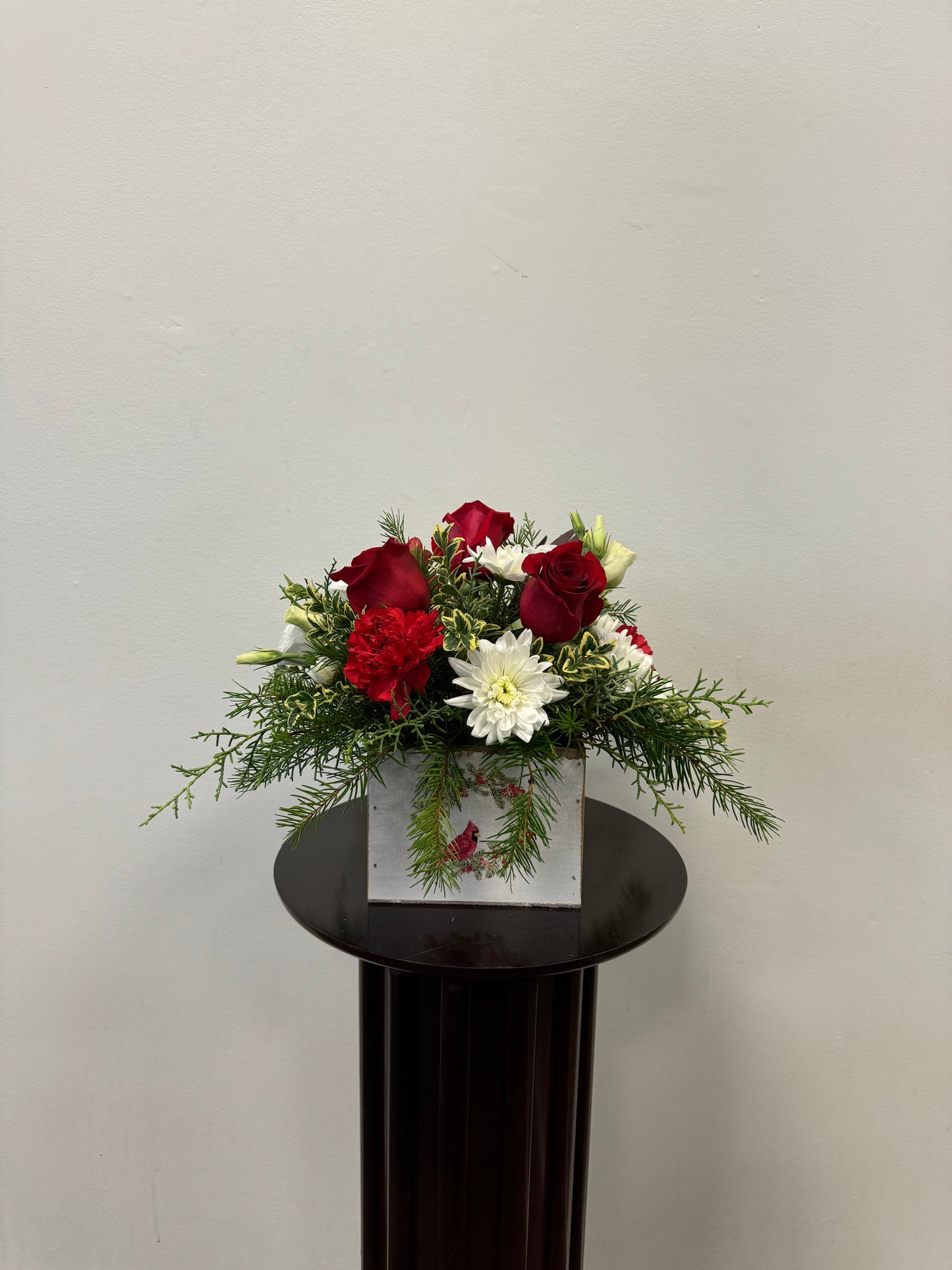 Introducing the Bird of Holiday Centrepiece from Posies Flowers & Fashion, showcasing a stunning arrangement of red roses, red carnations, white daisies, and lush greenery in an elegant glass vase. This beautiful display is set artfully on a dark pedestal against a light gray background.