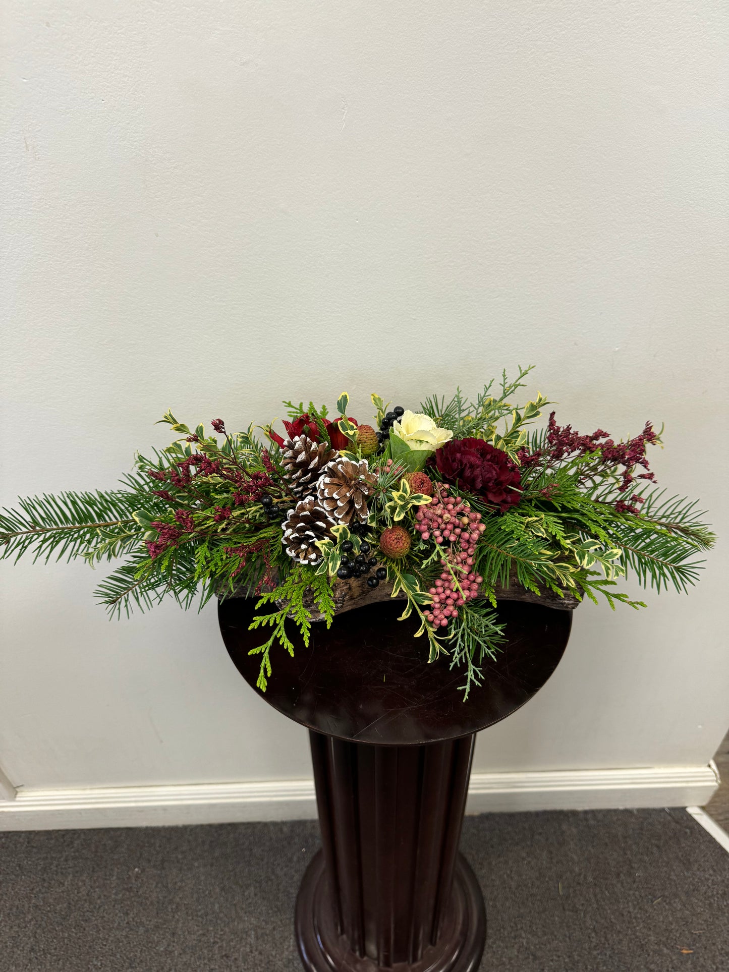 The Comfort of Home Centrepiece by Posies Flowers & Fashion exudes holiday cheer with an exquisite mix of fresh flowers, including pine cones, burgundy blooms, white roses, and pink berries. Enhanced by lush green foliage, it is beautifully displayed on a dark wood pedestal against a plain white wall.