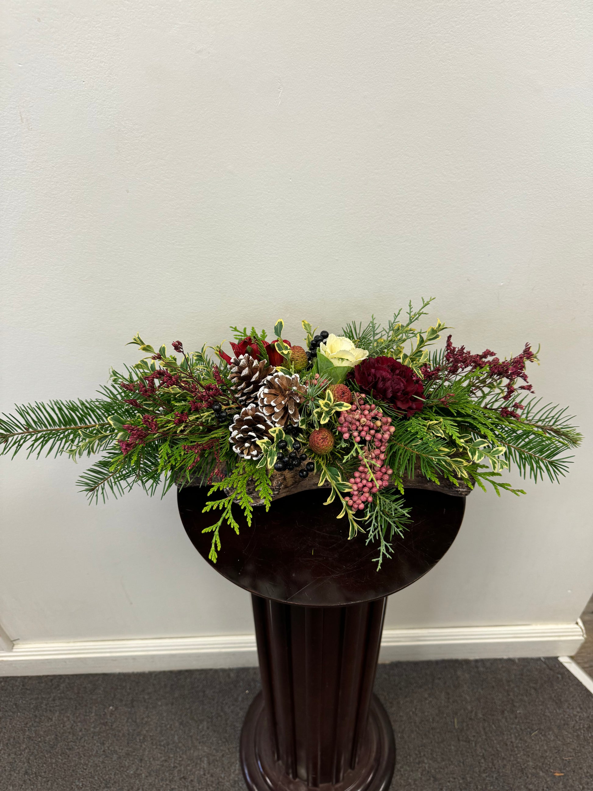 The Comfort of Home Centrepiece by Posies Flowers & Fashion exudes holiday cheer with an exquisite mix of fresh flowers, including pine cones, burgundy blooms, white roses, and pink berries. Enhanced by lush green foliage, it is beautifully displayed on a dark wood pedestal against a plain white wall.