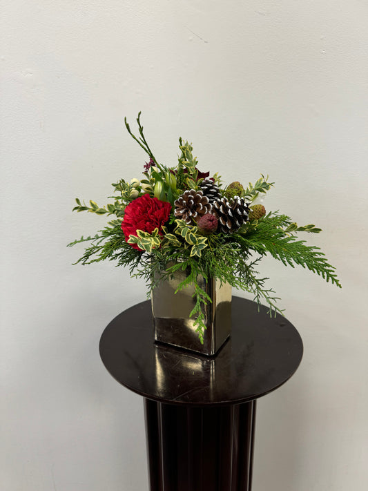 The Simple and Elegant Centrepiece by Posies Flowers & Fashion adds a festive charm to any room with its arrangement of red roses, greenery, pinecones, and small decorative elements in a square metallic vase. It's the perfect addition to a holiday table, elegantly displayed on a round dark pedestal against a plain white background.