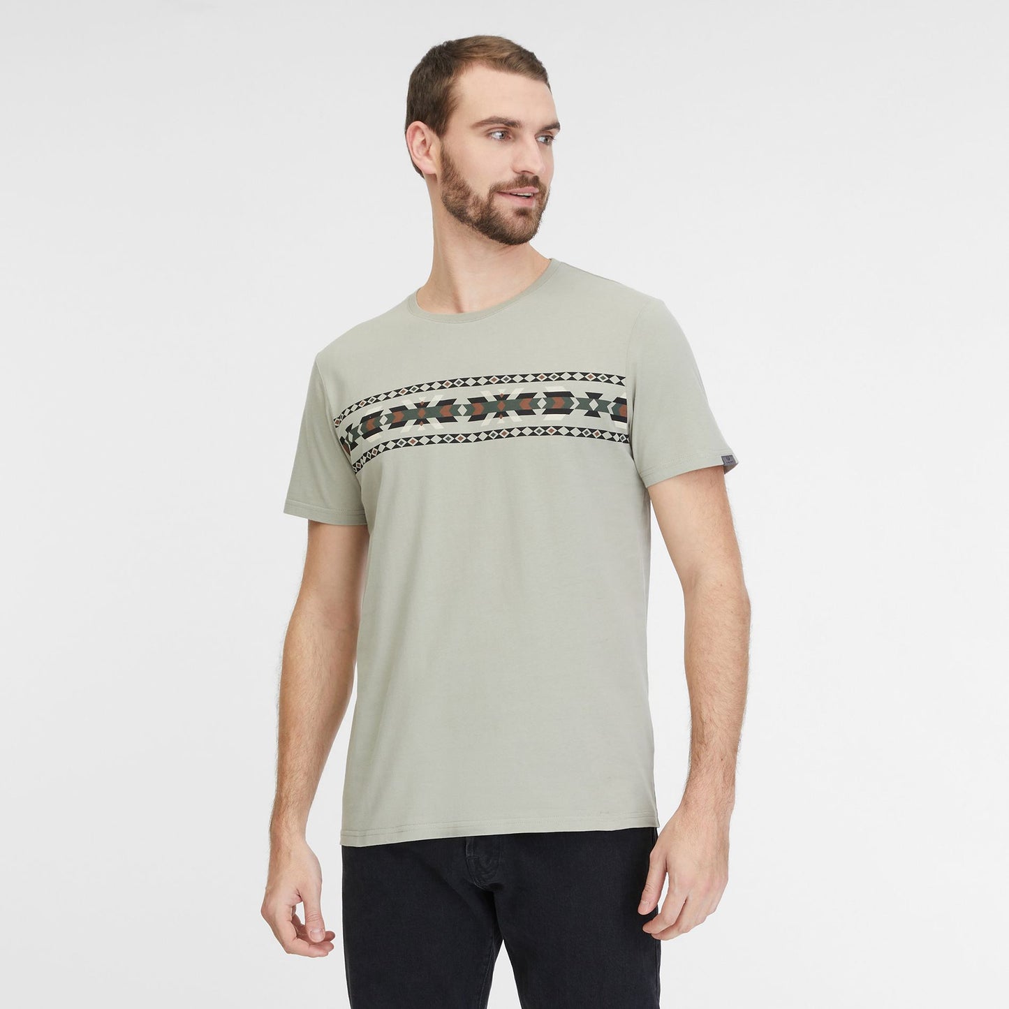 A man with short brown hair and a beard is wearing a Dusty Olive T-Shirt by RAGWEAR, made of 100% cotton and featuring a geometric pattern across the chest. He has black pants on and is standing against a plain white background, looking slightly to the right.