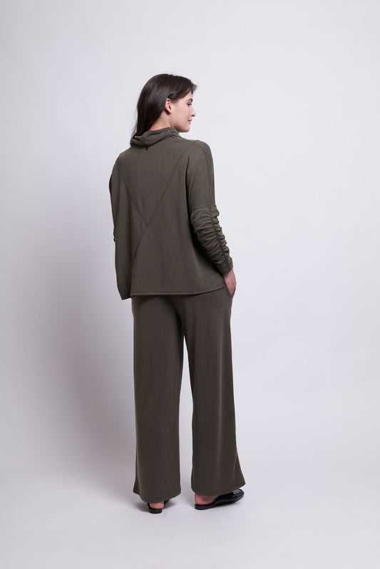 A person stands facing away, wearing an olive green, long-sleeved top paired with the So-Fa So Good Pant by FOIL. The versatile top features a cowl neck and textured detailing on the sleeves. The plain, light-colored background enhances the understated elegance of this matching wide-leg pant set.