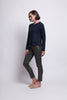 A person with short, dark hair stands against a plain white background. They are wearing a navy blue long-sleeve Raglan To Riches Sweater - FOIL made from recycled polyester, dark gray fitted pants, and beige slip-on shoes. They are looking down and slightly to their left with their arms relaxed by their sides.