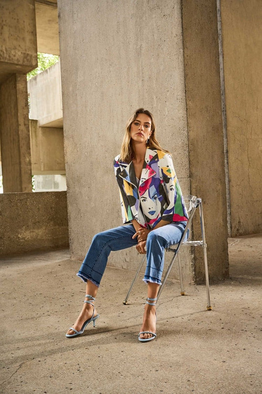 Against a concrete backdrop, a woman sits on a metal chair wearing Joseph Ribkoff’s artistic print jacket, Flare Crop Stretch Jeans 251957 with frayed hem by Joseph Ribkoff, and strappy heels. Her expression is contemplative in the stylish industrial setting.