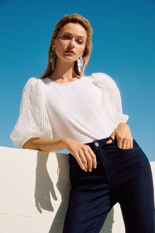 A person with long hair poses against a clear blue sky, wearing the Jersey Straight Top With Novelty Sleeves by Joseph Ribkoff, dark jeans, and large statement earrings.