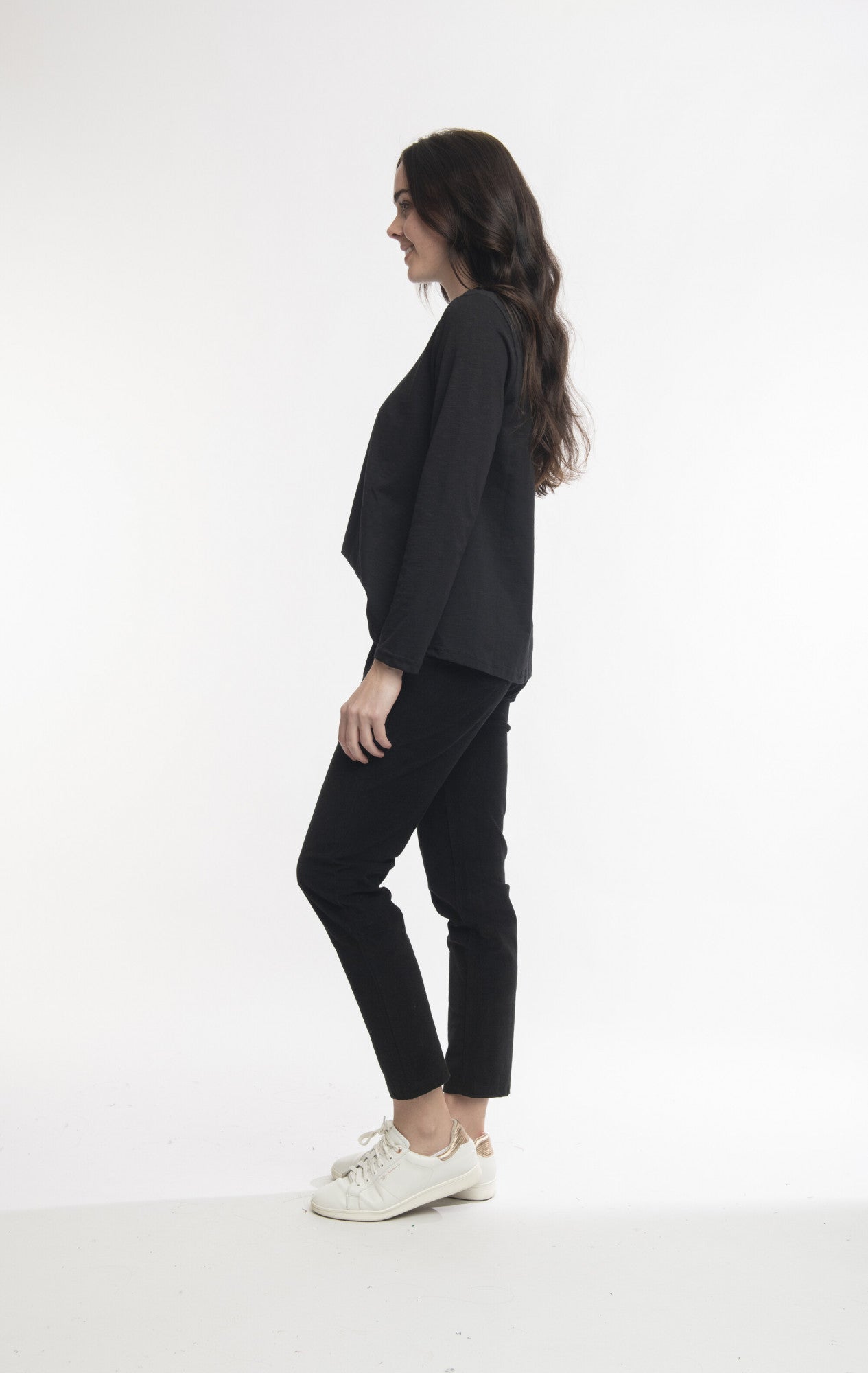 A person with long, wavy hair stands facing left against a white background. They are wearing a black Jumana Jacket made of durable polyester, paired with Solid Cord Jeggings 2612 by ORIENTIQUE and white sneakers. The individual has their hands relaxed at their sides and is captured in a standing pose.