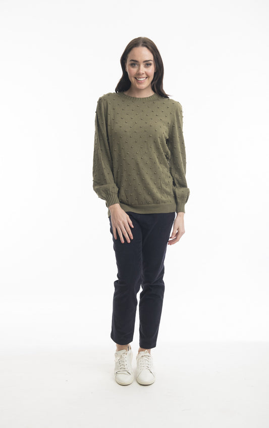 A woman with long dark hair stands smiling, dressed in an ORIENTIQUE Bobble Knit Jumper (Style: 1255), a green textured long-sleeve shirt with polka dot detailing. She pairs it with dark pants and white sneakers against a plain white background.