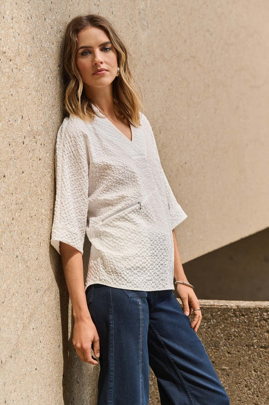 A woman with shoulder-length hair leans against a textured wall, wearing the Joseph Ribkoff Seersucker Boxy High-Low Top 251165 and blue jeans, looking attentively at the camera. The neutral-toned background complements her casual outfit effortlessly.