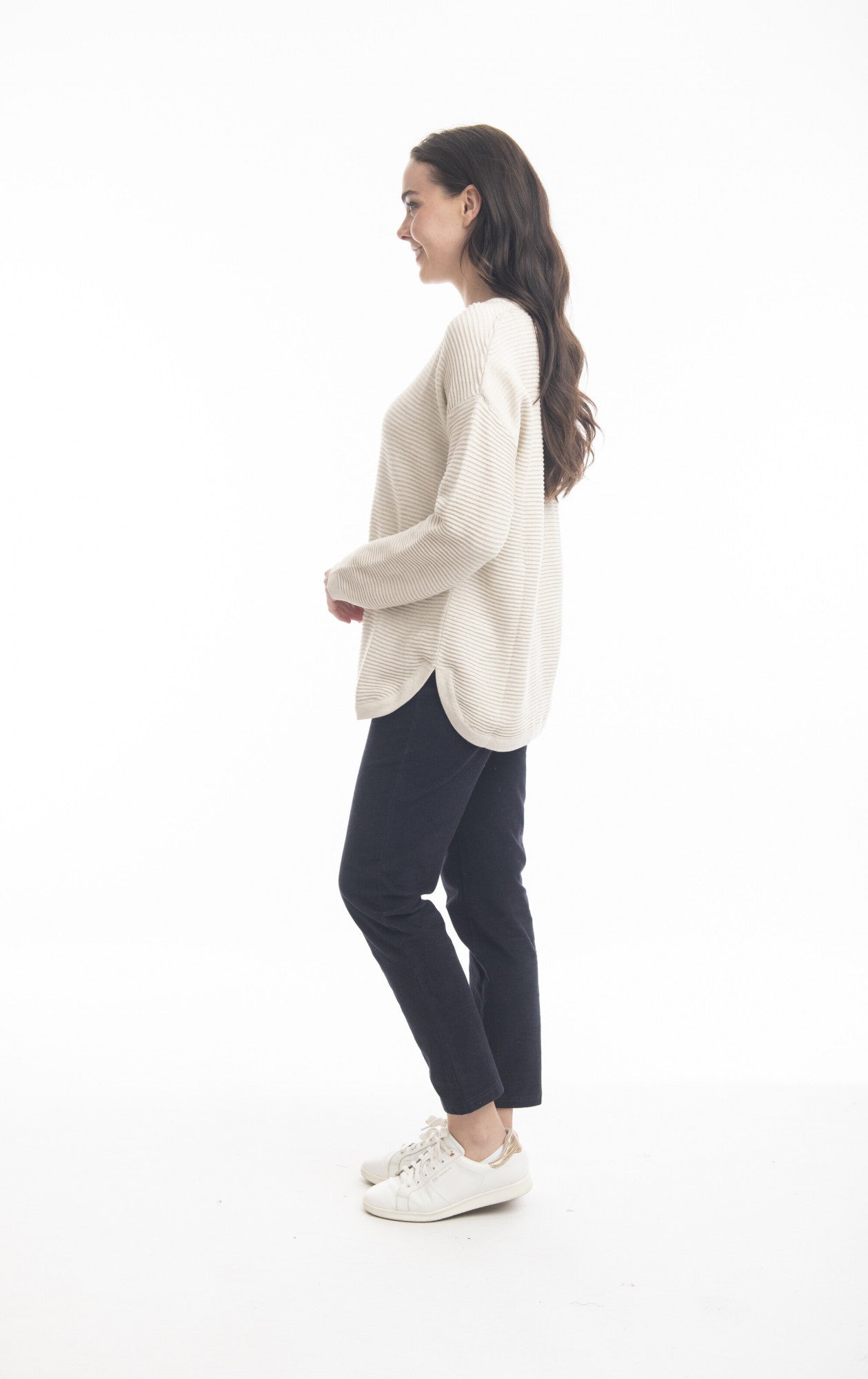 A woman with long brown hair stands in a side profile against a white background. She is wearing the Ribbed Knit Jumper Style: 1253 by ORIENTIQUE, paired with black pants and white sneakers. She's smiling and appears relaxed, effortlessly showcasing sustainable fashion.