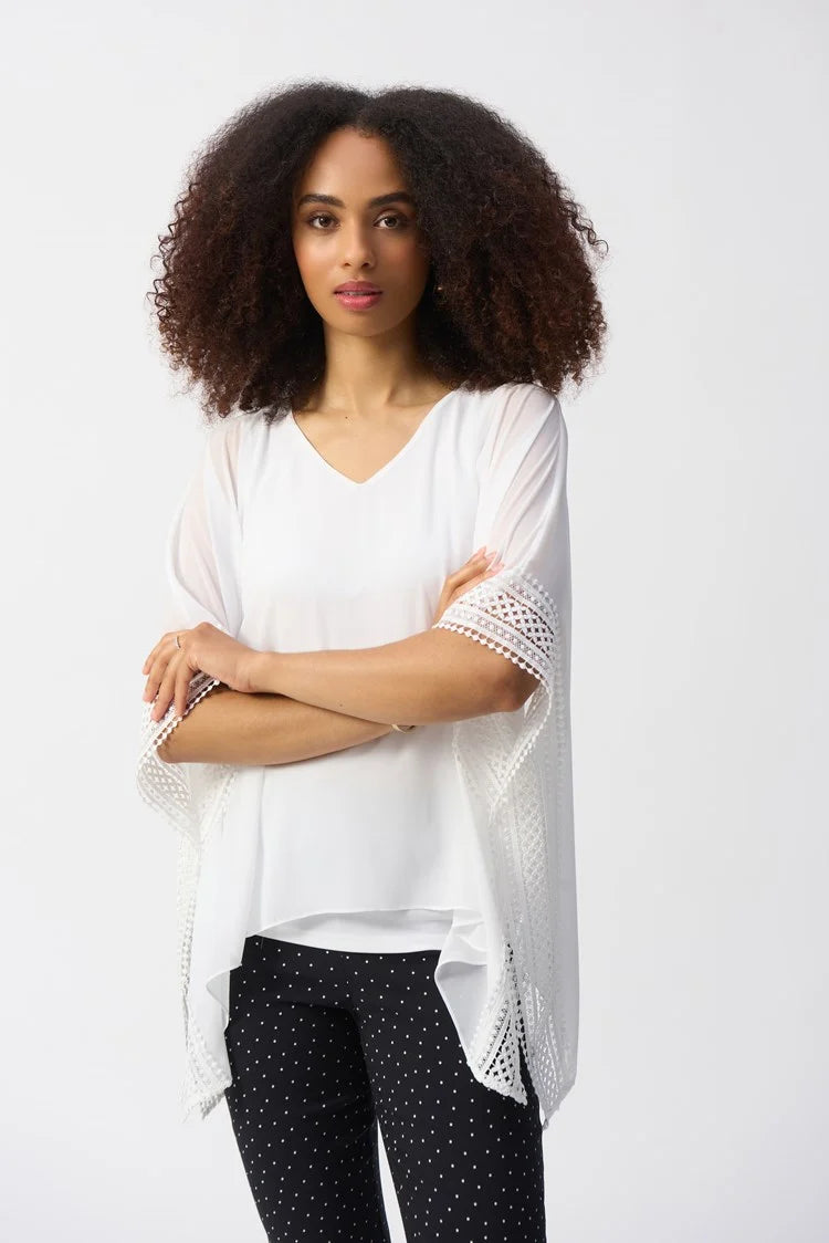 A curly-haired woman wears the Joseph Ribkoff Chiffon And Silky Knit Poncho Top 251095 with a V-neckline and lace-detailed sleeves, paired with black polka-dot pants. She stands against a plain background, arms crossed, looking directly at the camera.