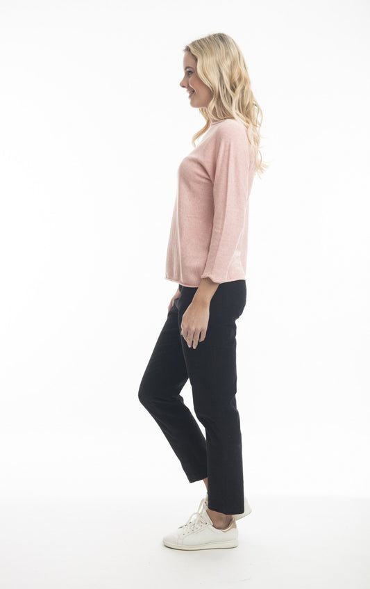 A woman stands in profile against a white background. She has blonde hair and is wearing a light pink, 100% organic cotton Boat Neck Knit Top with Long Sleeve by ORIENTIQUE (Style: 1259 - Orientique), black pants, and white sneakers. She appears relaxed and is smiling slightly.