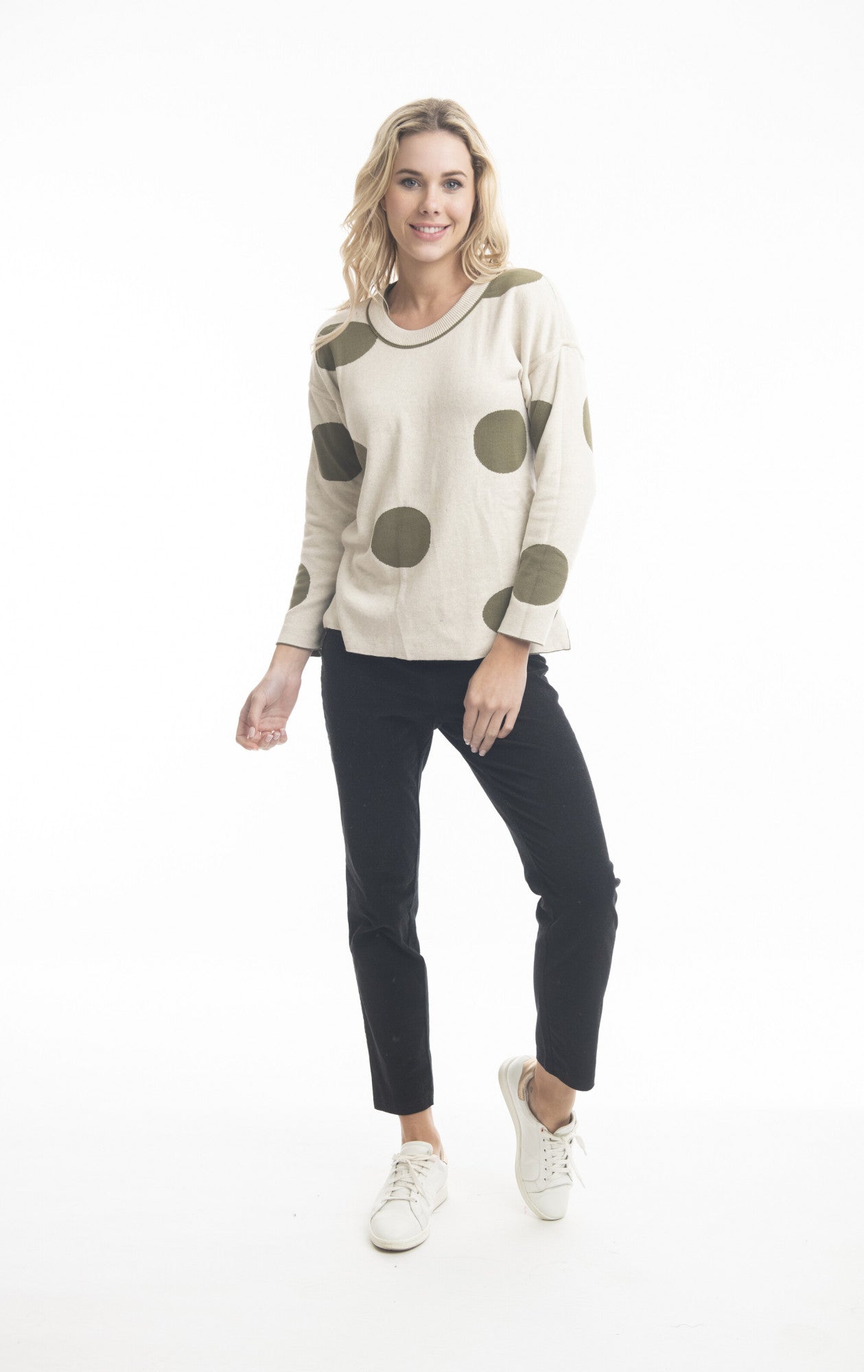 A person with long, wavy blond hair is wearing a beige Reversible Knit Jumper Style: 1267 - Orientique from ORIENTIQUE, which features large green polka dots. They are also dressed in black pants and white sneakers. The fun print of their 100% organic cotton jumper stands out as they pose against a plain white background, smiling and standing with one foot slightly raised.