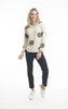 A person with long, wavy blond hair is wearing a beige Reversible Knit Jumper Style: 1267 - Orientique from ORIENTIQUE, which features large green polka dots. They are also dressed in black pants and white sneakers. The fun print of their 100% organic cotton jumper stands out as they pose against a plain white background, smiling and standing with one foot slightly raised.