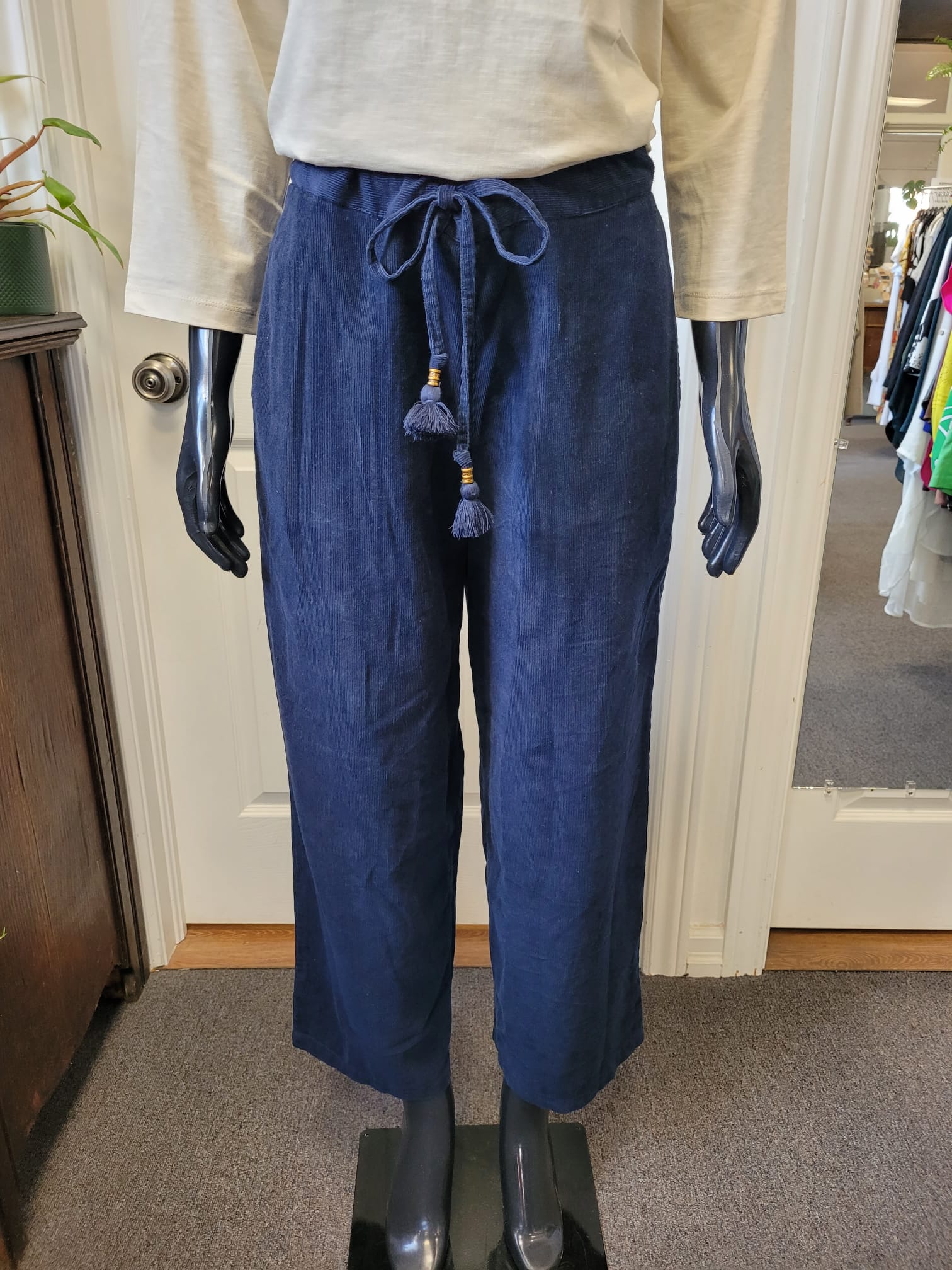 A mannequin dressed in a beige long-sleeve top and ORIENTIQUE's navy blue, high-waisted Cord Solid Trousers Flared Style: 2611, featuring a waist tie and tassel detail. The mannequin stands on a carpeted floor next to a wooden cabinet and an open door with a clothes rack visible in the background.