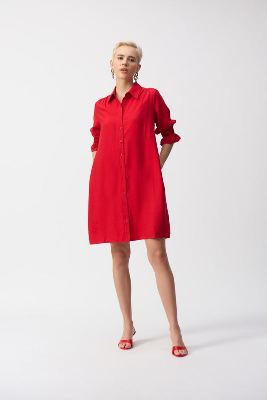 A person confidently poses against a white background wearing the Woven Button-Front Shirt Dress 251947 by Joseph Ribkoff, styled in vibrant red with button-down detailing and pockets, matched with red heels. Their short, blonde hair complements the ensemble.