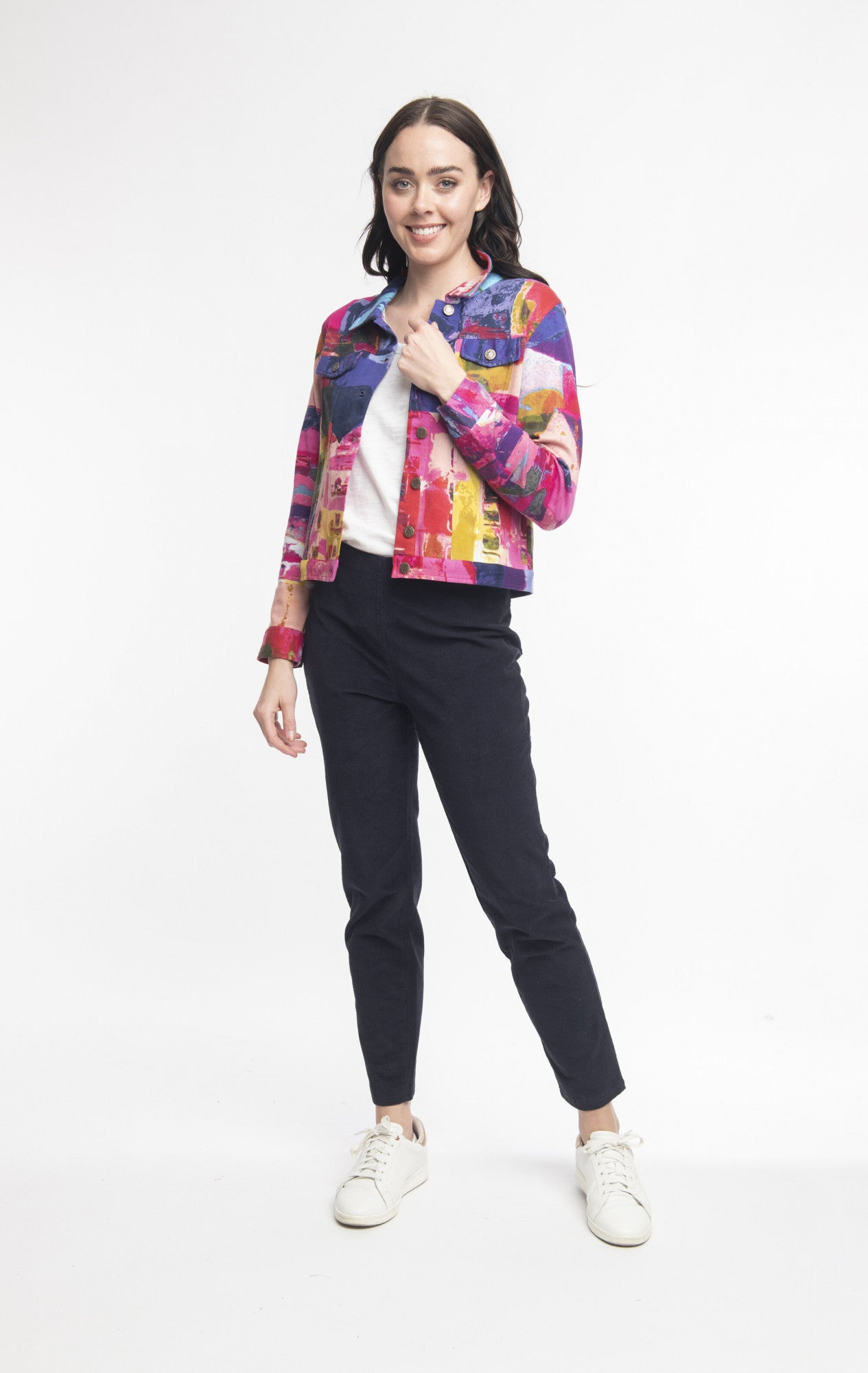 A woman stands against a plain white background, smiling. She is wearing the colorful, patterned Biker Jacket Style: 22907 by Joseph Ribkoff from ORIENTIQUE over a white top, black pants, and white sneakers. With shoulder-length brown hair and vibrant colors brightening her outfit, she poses with one hand on her hip and the other by her side.