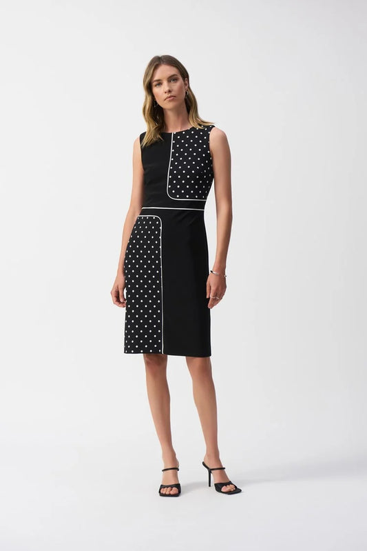 A woman models the Joseph Ribkoff Silky Knit Dot Print Sleeveless Sheath Dress, 251094. It features a black sheath silhouette adorned with white polka dots, solid black sections, and elegant white trim. She completes the look with black heels against a simple white backdrop.