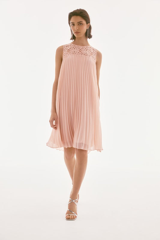 A person models the Joseph Ribkoff Signature Chiffon and Lace Sleeveless Pleated Dress 251767, a knee-length pink trapeze dress featuring a lace yoke. They stand gracefully with one foot forward against a simple, light background.