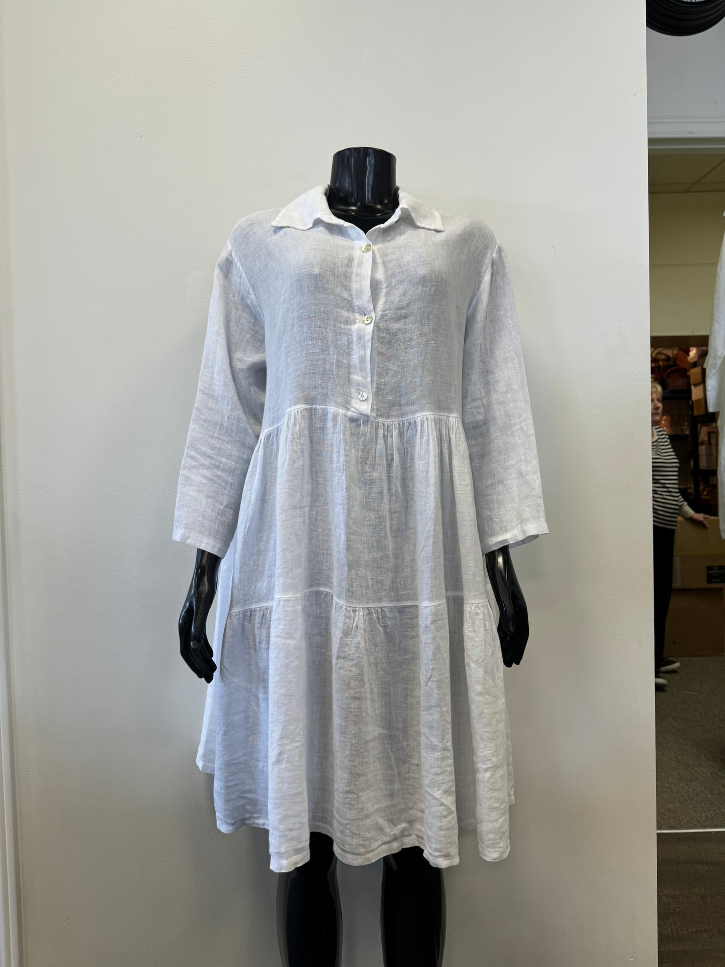 A mannequin showcases the Linen Dress - WHITE Catherine Lillywhite by Catherine Lillywhite against a plain, light-colored background. The airy summer essential is 100% linen and features long sleeves, a collar, and buttons down the top half, with a charming tiered skirt adding to its loose-fitting elegance.