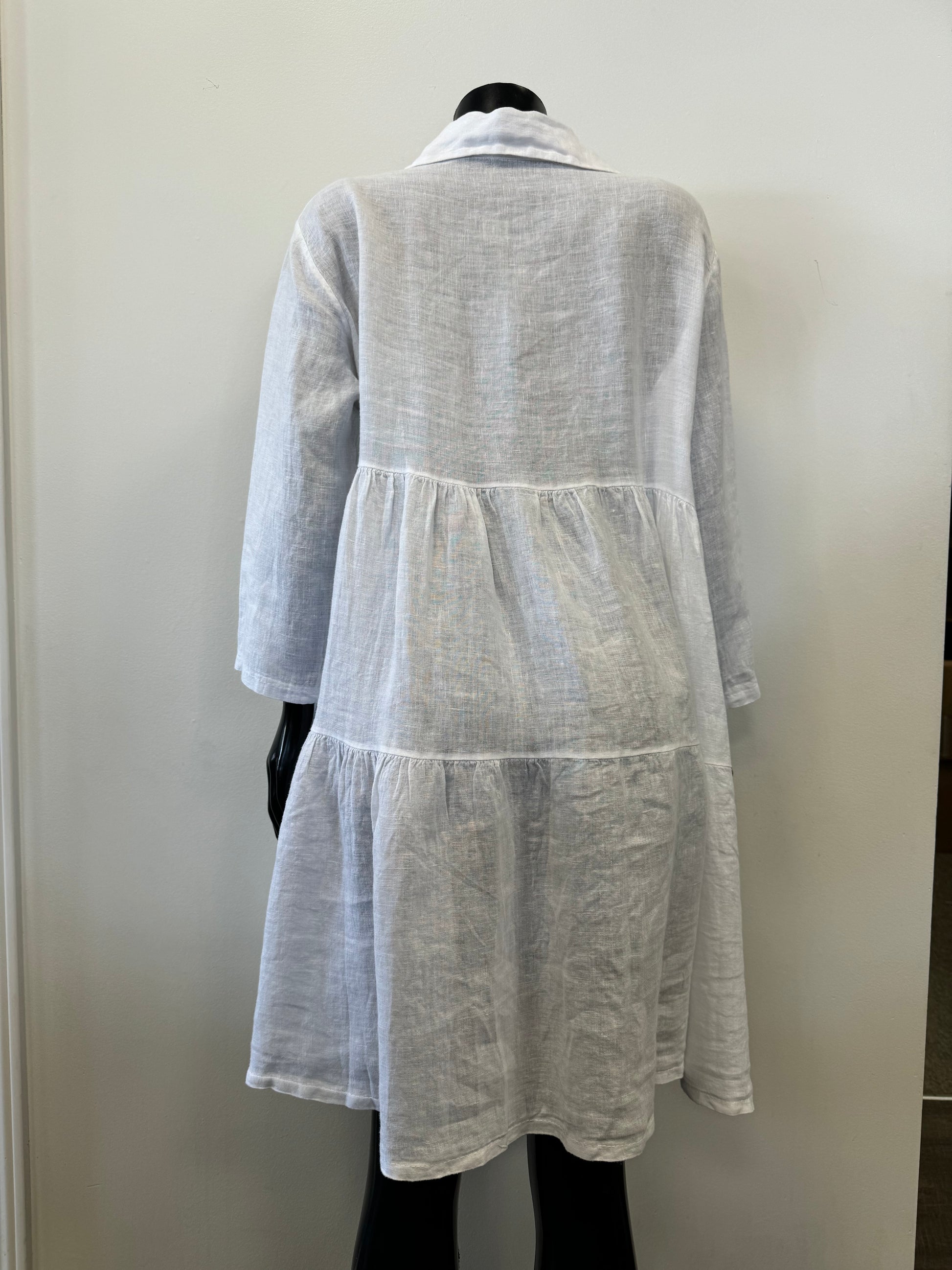 A mannequin showcases the Linen Dress - WHITE Catherine Lillywhite by Catherine Lillywhite against a plain, light-colored background. The airy summer essential is 100% linen and features long sleeves, a collar, and buttons down the top half, with a charming tiered skirt adding to its loose-fitting elegance.